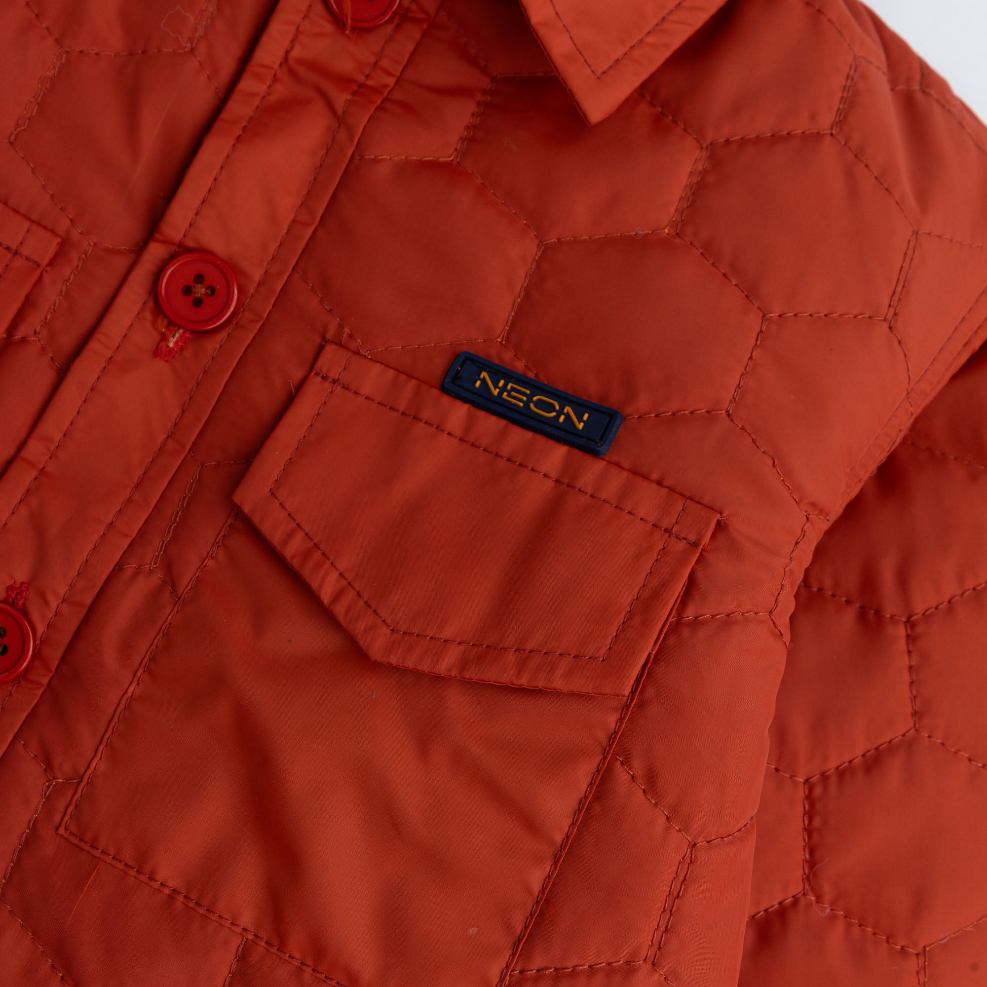 Close-up view of the rust parachute fabric, hexagonal quilted stitching, chest pocket and NEON logo detail on a boys shirt jacket.