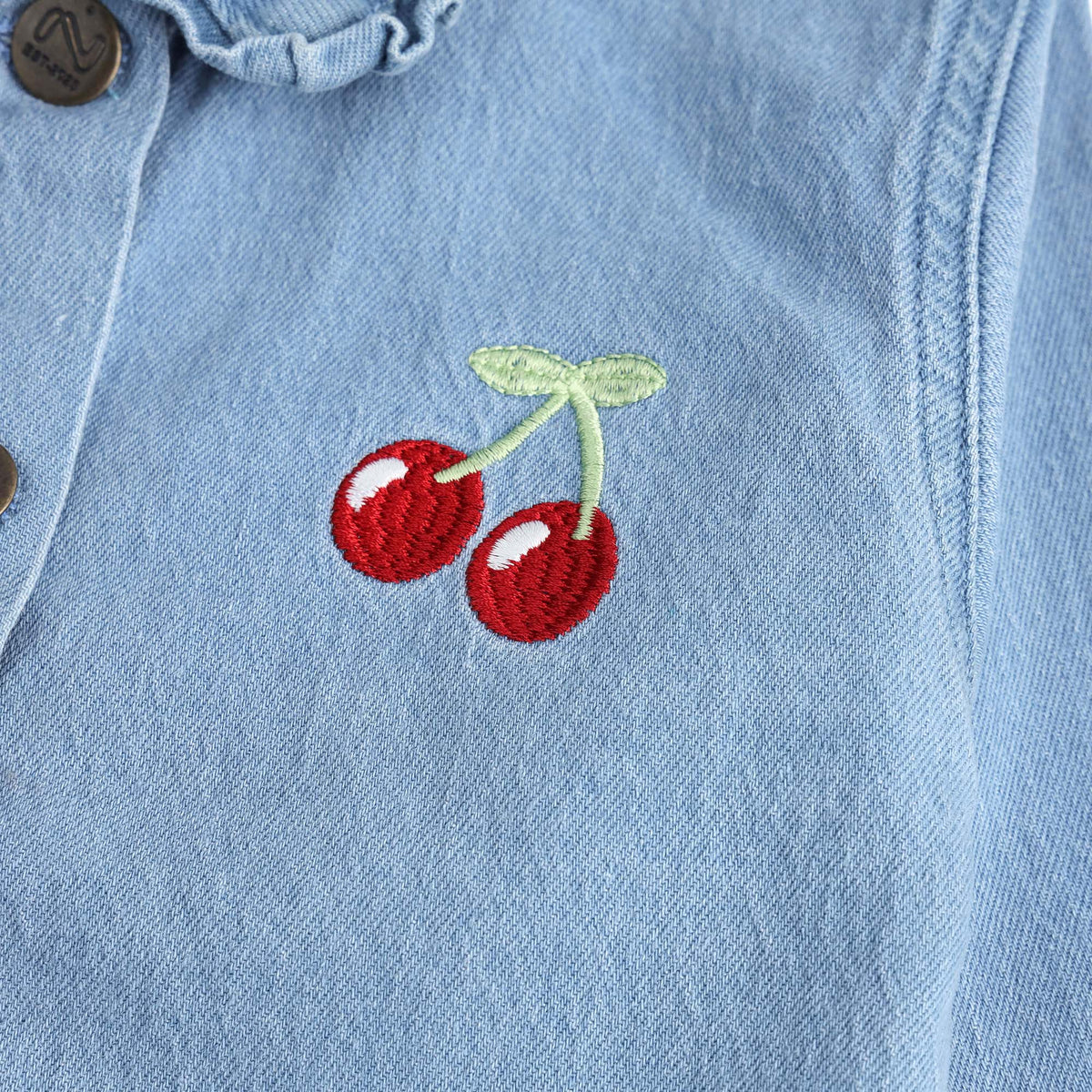 Close-up view of the red cherry embroidery with mint-green stems and white highlights on the girls light-wash denim jacket.