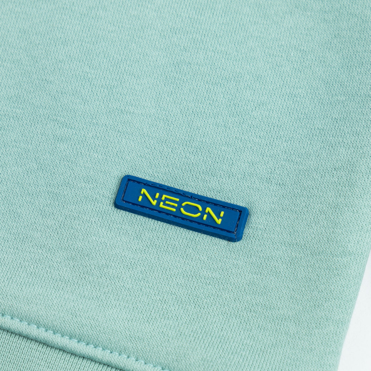 Close-up of the navy blue and neon yellow NEON logo patch on the kids mint green fleece sweatshirt.