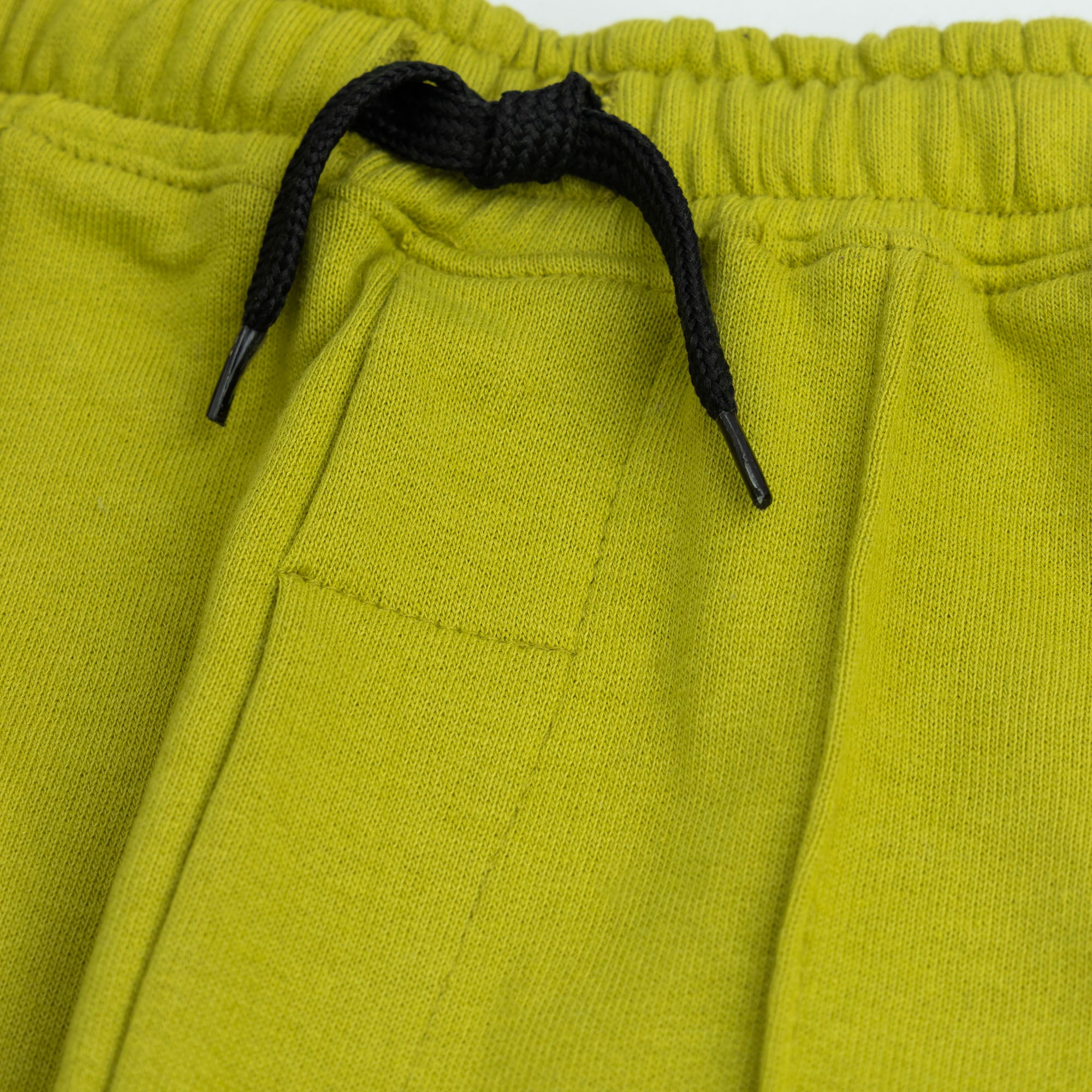 Close-up of the elasticated waistband, contrast black drawstring and front pintuck seams on the boys’ lime green terry joggers.