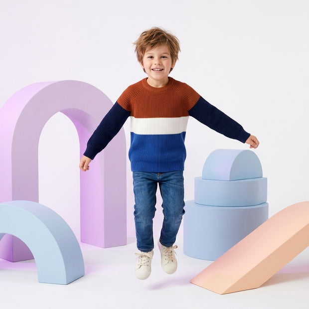 Young boy wearing a rust, navy, white and blue color-block sweater paired with blue denim.
