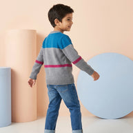Back view of a young boy wearing a grey color-block sweater with blue and maroon panels, paired with blue denim.
