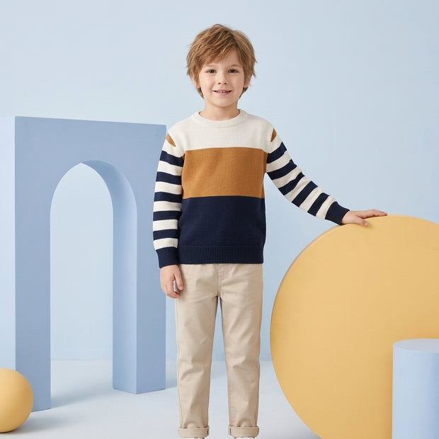 Young boy wearing a white, brown and navy color-block sweater with striped sleeves, paired with beige trousers.