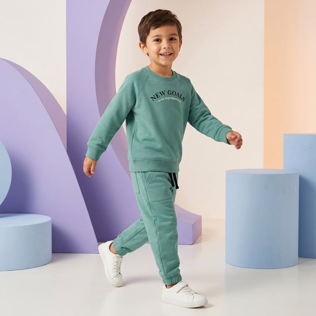 Young boy wearing a sage green New Goals fleece co-ord set featuring a graphic sweatshirt and matching joggers.