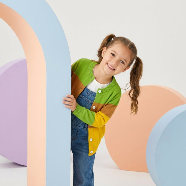 Young girl wearing a green, brown and yellow color-block button cardigan paired with blue denim overalls.