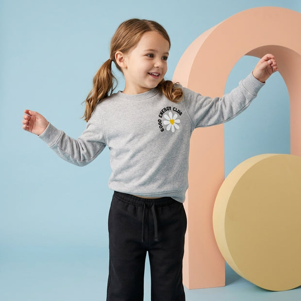 Smiling young girl wearing a grey daisy graphic fleece sweatshirt paired with black drawstring trousers.