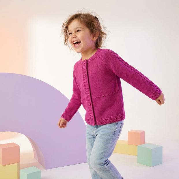 Smiling young girl wearing a deep pink button-front cardigan with cable-pattern sleeves, paired with light-wash denim.