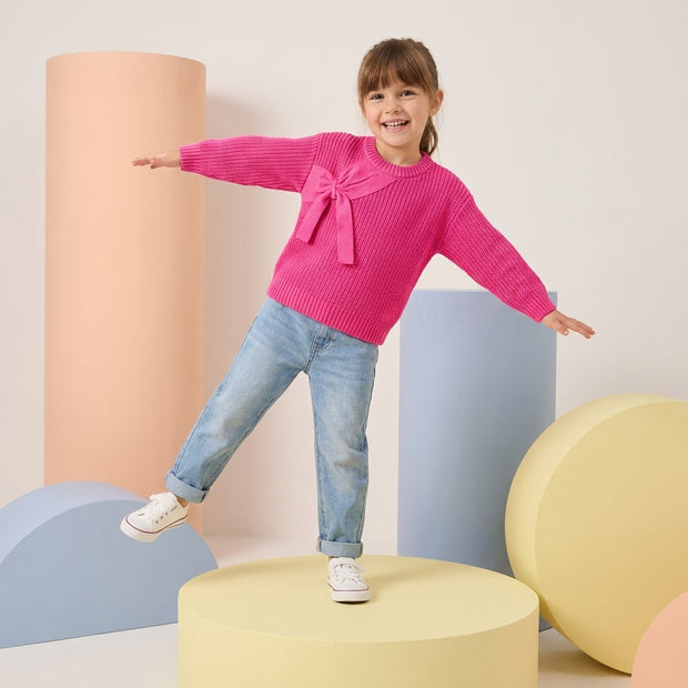 Smiling young girl wearing a bright pink sweater with an oversized front bow and light-wash denim.