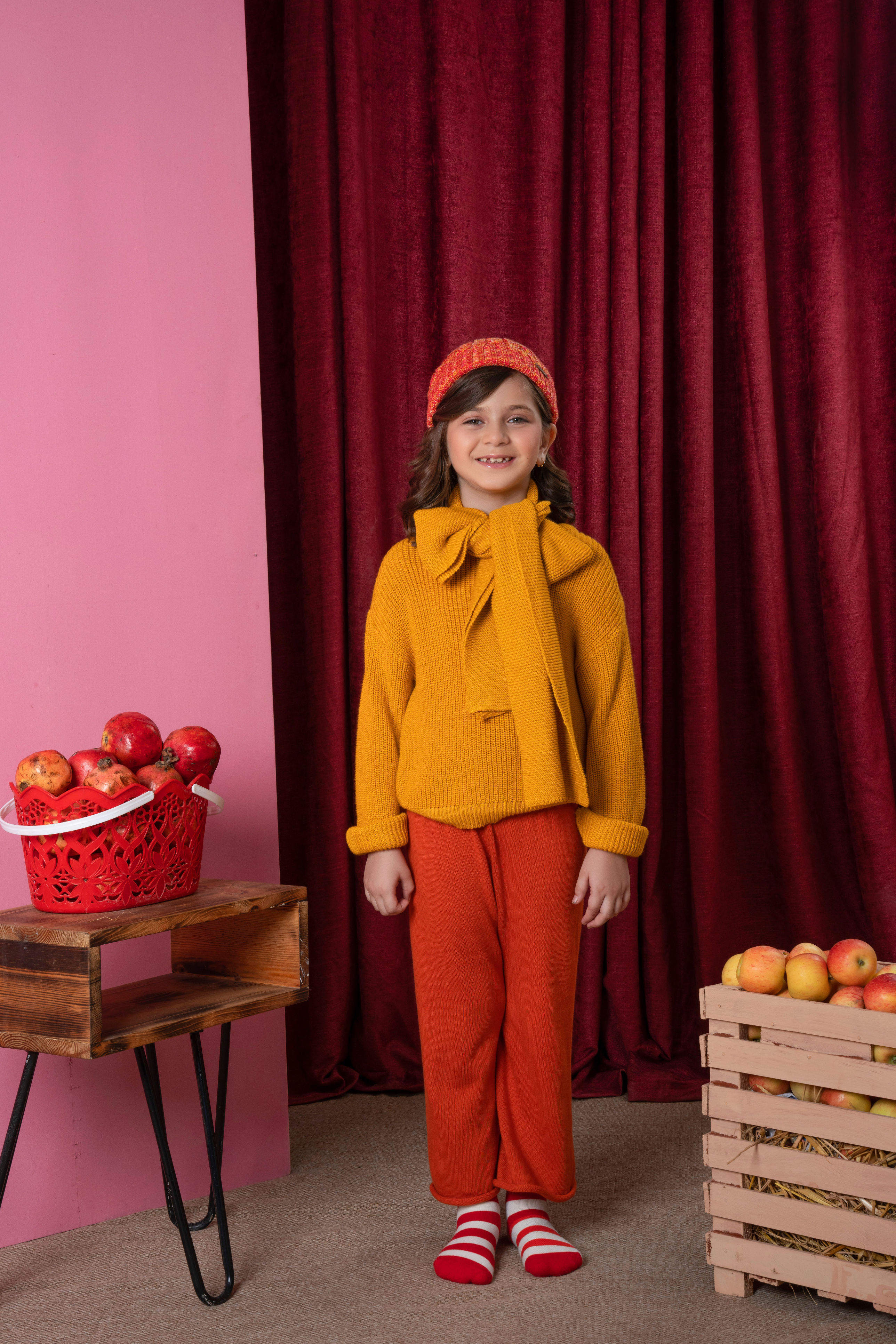 Girl wearing a mustard knitted top and rust orange trouser co-ord set with a matching headband in a colorful indoor winter setting.